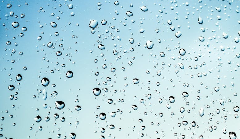 A piece of clear glass is covered with droplets of pure water with a light blue sky beyond.