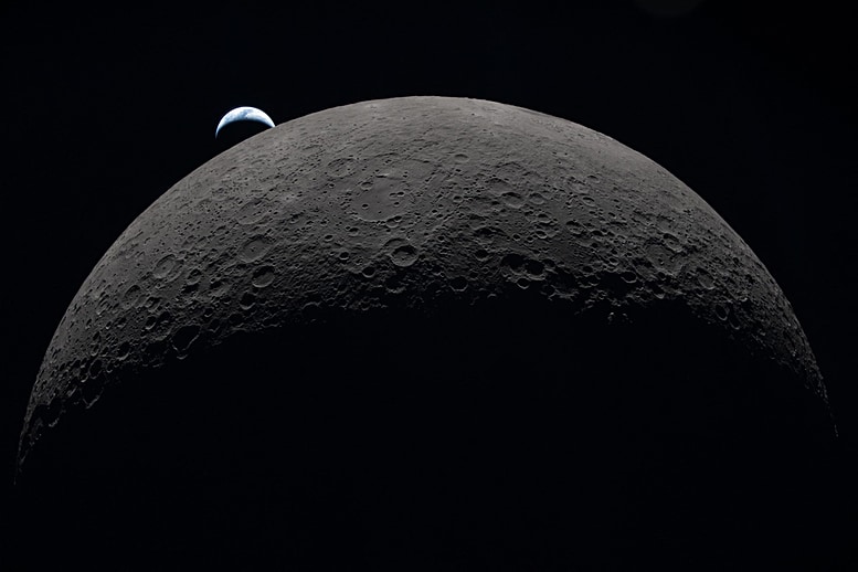 A view of the dark side of the moon. Only the top half of it can be seen, dark gray in color, floating in the blackness of space. Floating beyond it, just above the top left portion of the moon, is a crescent of a tiny blue-white marble, our Earth, just as it is about to set behind the moon.